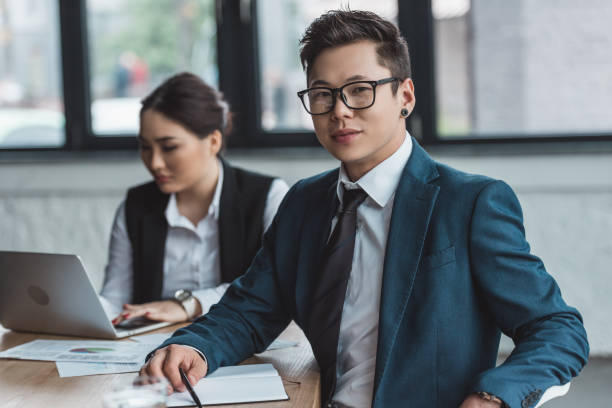 istock handsome young asian businessman in eyeglasses smiling at camera while female colleague using laptop behind in office 1089367254