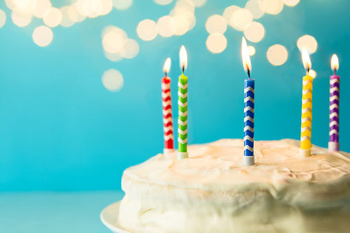 White birthday cake with colorful candles on blue background against defocused light. Holiday celebration concept. Copy space