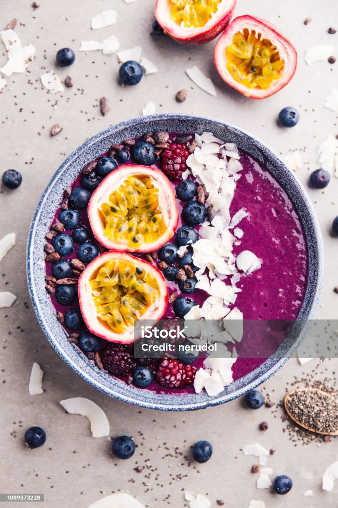 Smoothie acai bowl served in bowl on grey table Tasty appetizing smoothie acai bowl made from blackberries and wild berries, decorated with cut passion fruit, coconut flakes, and cacao nibs. Served in bowl. Healthy life clean eating concept. Bowl Stock Photo