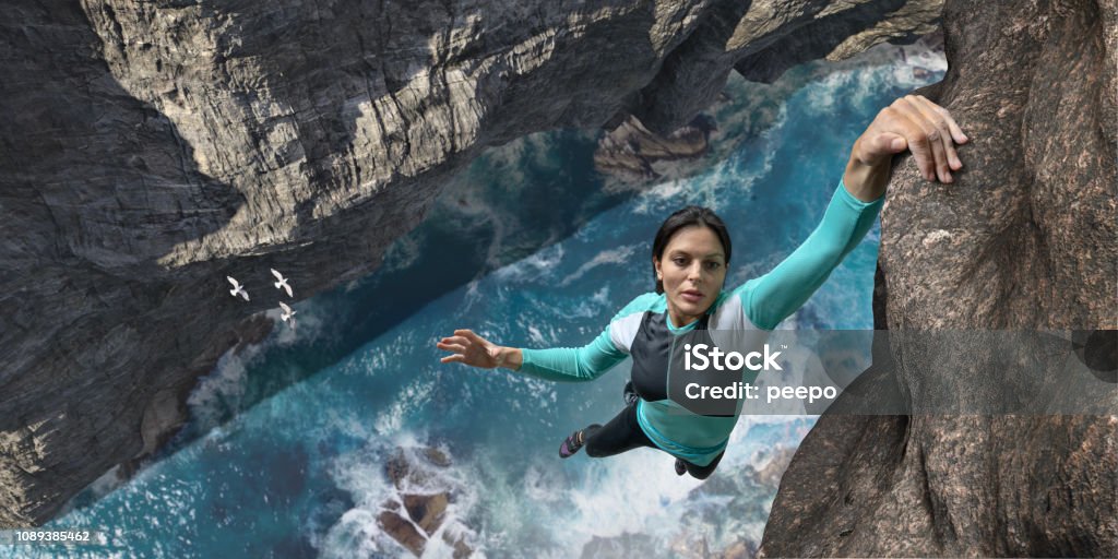 Free Climber Hangs One Handed On Sea Cliff Rock Face A young woman extreme free climbing without safety equipment, hangs one handed from a rock face over the sea and rocks below. The climber is wearing climbing top, leggings. chalk bag and climbing shoes. Waves break over rocks in the sea below during sunny daylight in clear weather. Women Stock Photo