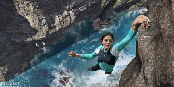 Free Climber Hangs One Handed On Sea Cliff Rock Face A young woman extreme free climbing without safety equipment, hangs one handed from a rock face over the sea and rocks below. The climber is wearing climbing top, leggings. chalk bag and climbing shoes. Waves break over rocks in the sea below during sunny daylight in clear weather. extreme sports stock pictures, royalty-free photos & images
