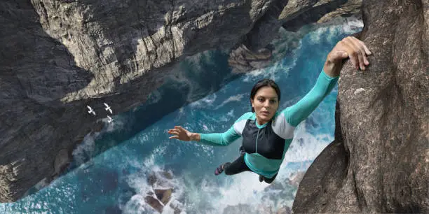 A young woman extreme free climbing without safety equipment, hangs one handed from a rock face over the sea and rocks below. The climber is wearing climbing top, leggings. chalk bag and climbing shoes. Waves break over rocks in the sea below during sunny daylight in clear weather.