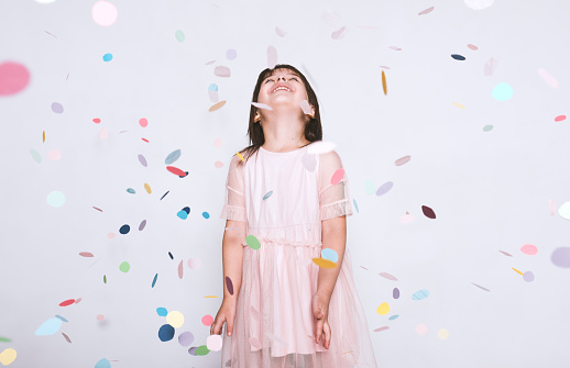 Beautiful little girl wearing pink dress in tulle with princess crown looking up to the colorful confetti surprise posing on white studio wall. Happy girl celebrating her birthday party, having fun