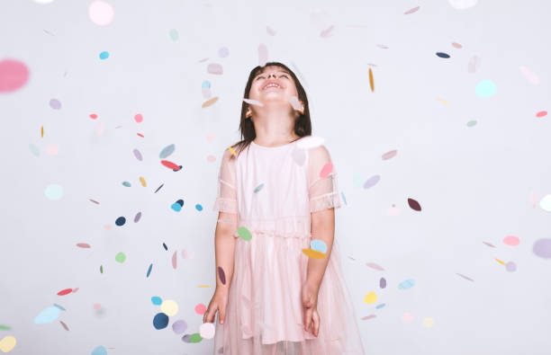 Beautiful little girl wearing pink dress in tulle with princess crown looking up to the colorful confetti surprise posing on white studio wall. Happy girl celebrating her birthday party, having fun stock photo