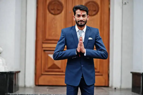 Stylish beard indian man with bindi on forehead, wear on blue suit posed outdoor against door of building and show namaste hands sign.
