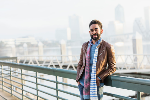 A stylish mixed race African-American and Hispanic mid adult man in his 30s standing on a city waterfront, wearing a leather jacket, sweater and scarf, smiling confidently at the camera.