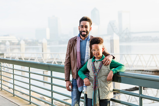 A mixed race African-American and Hispanic mid adult man in his 30s standing with his teenage son, 13 years old, on a city waterfront, smiling at the camera.