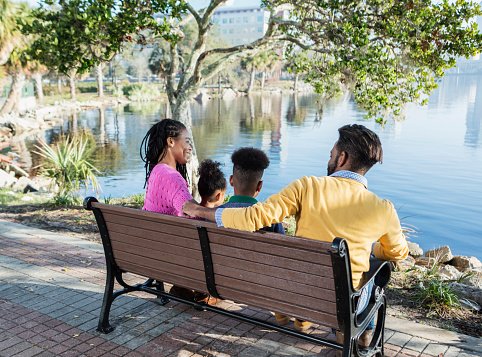 Rear view of a mixed race family sitting on a park bench overlooking a city waterfront, with their two children, a 13 year old teenage boy and 3 year old daughter. The mother is African-American and father is African-American and Hispanic.