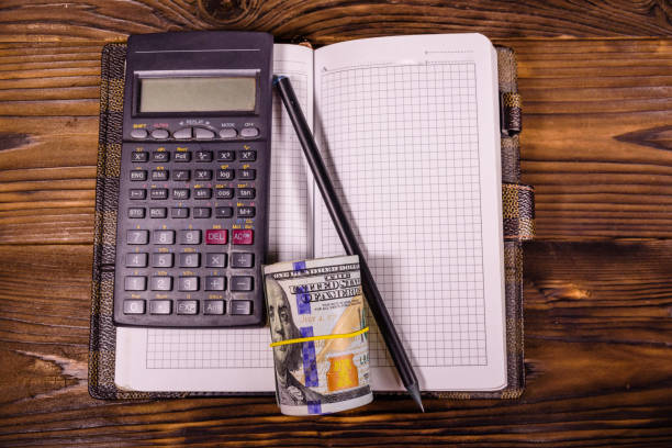 istock Opened notepad, one hundred dollar banknotes, scientific calculator and pencil on wooden table. Top view 1089407454