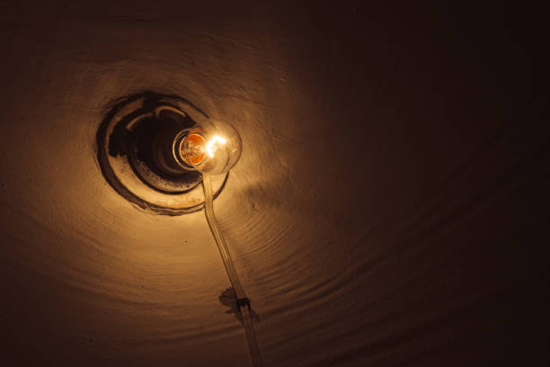 Incandescent lamp burns inside dark room close-up. Illumination with copy space. Old light bulb on rough wall. Lamp illuminate basement. Light in darkness. Minimalist background. Warm orange lighting.