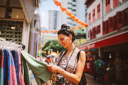Vintage toned street style portrait of a young fashionable brunette woman, walking down the busy streets of Singapore Chinatown district, shopping through the market stalls. She is wearing a summertime dress, holding her analog SLR camera, or maybe a new mirrorless camera in a fashionable retro body.