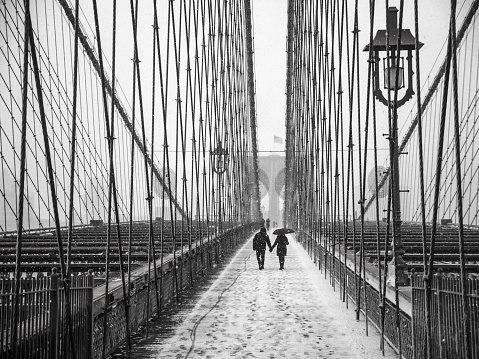 A snowy scene of people with umbrellas in silhouette walking across the Brooklyn Bridge from Manhattan to Brooklyn.