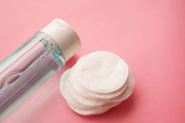 Photo of bottle with tonic or micellar cleansing water with fresh herbs and flowers and cotton pads on pink background, top view. Beauty, skin, hair or body care concept. selective focus