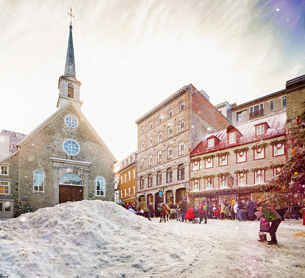 Quebec City Christmas Winter scene at an icy Place Royale with light snow fall, featuring the Notre-Dame des victoires church and a crowd of people walking by shops and cafes.
