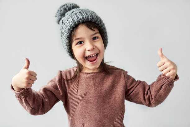 Horizontal closeup portrait of happy smiling little girl in the winter warm gray hat, wearing sweater and  showing thumbs up, posing on a white studio background. stock photo