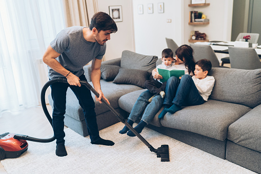 Man vacuuming while mother and boys reading book on the sofa