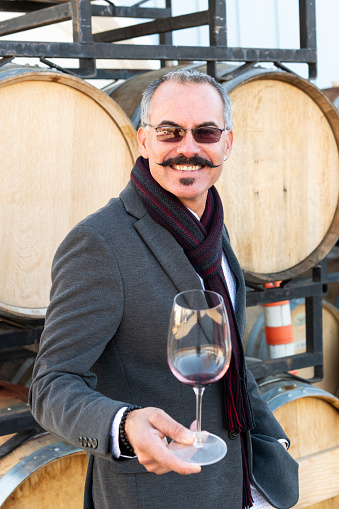 mature man posing holding a glass of red wine with wine barrels in the background