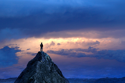 A male hiker stands on top of a mountain peak and looks at the sunset in the distance.