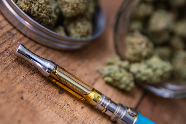 THC Oil Concentrate Filled Vape Pen Up Close THC oil concentrate filled vape pen on natural wood with an open glass container full of Mango Kush strain marijuana buds grown & sold in dispensaries through-out Southern California. cannabis store photos stock pictures, royalty-free photos & images