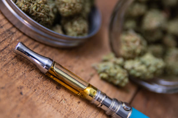 THC Oil Concentrate Filled Vape Pen Up Close THC oil concentrate filled vape pen on natural wood with an open glass container full of Mango Kush strain marijuana buds grown & sold in dispensaries through-out Southern California. cannabaceae stock pictures, royalty-free photos & images