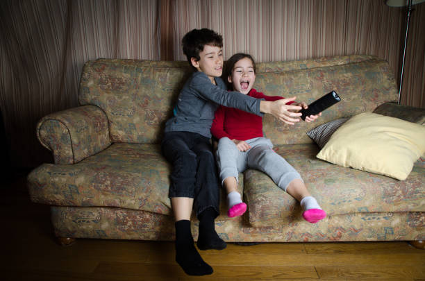 двое детей борются за пульт дистанционного управления телевизором, сидя на диване в гостиной - family television fighting watching стоковые фото и изображения