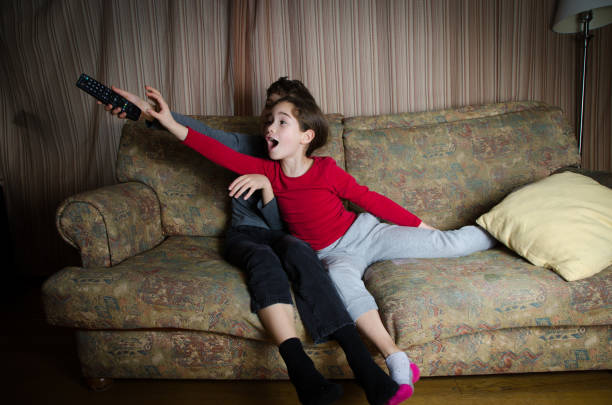 двое детей борются за пульт дистанционного управления телевизором, сидя на диване в гостиной - family television fighting watching стоковые фото и изображения