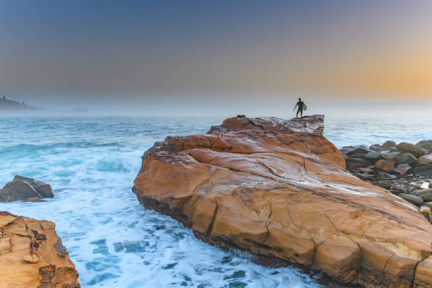 Early Morning Misty Seascape with Rock and Surfer Capturing the sunrise from Avoca Beach on the Central Coast, NSW, Australia. avoca beach photos stock pictures, royalty-free photos & images
