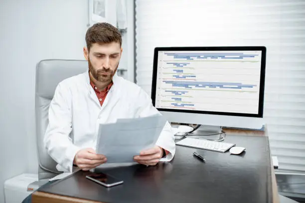 Handsome doctor reading some documents sitting in the luxury medical office with computer on the background