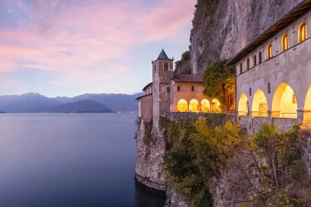 The Hermitage of Saint Catherine of stone (eremo di Santa Caterina del Sasso) in Leggiuno, is a tourist destination and itinerary on Lake Maggiore in province of Varese and in northern Italy. Ancient religious building and evocative sunset. In the background the lights of the cities of Intra and Pallanza