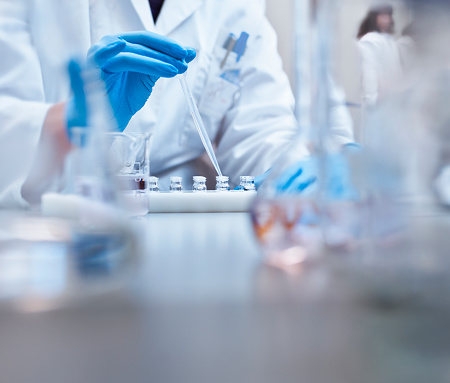 Scientist filling vials with solution through pipette in test tube rack. Chemist is examining medicine during scientific experiment. She is wearing gloves at pharmaceutical factory.