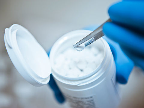 Cropped image of scientist holding medicine with tweezers. Pills are in white bottle. She is wearing gloves at pharmaceutical factory.