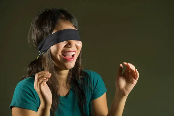 Photo of young happy and cute blindfolded Asian Korean teenager girl excited playing dangerous internet viral challenge isolated on dark background under edgy and dramatic studio light