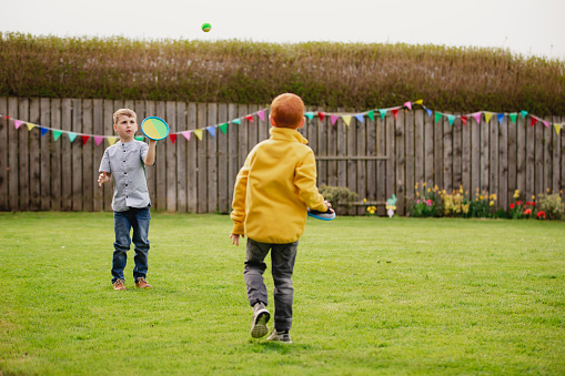 Two young boys playing outside in a back garden. They are throwing a tennis ball to each other and catching it with a velcro mitt.