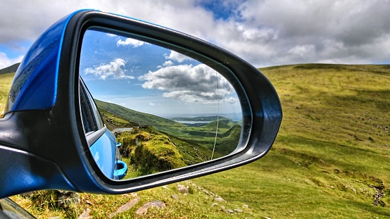 Looking through a rearview mirror at the car on a panorama of a landscape with lush green fields and meadows. In the background you have a fantastic view of rocks, island and sea. The landscape is located in Ireland