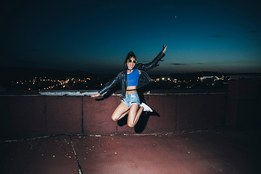 Cheerful young woman having fun on roof balcony, jumping, at night.