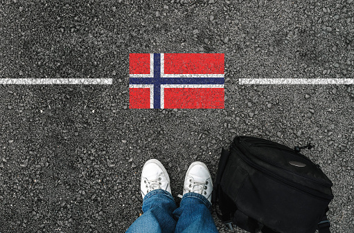 a man with a shoes and backpack is standing on asphalt next to flag of Norway and border