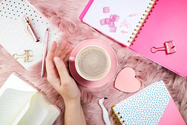 Photo of Flat lay femininity background. Female hand, coffee cup, notebooks and office supplies on pink woolen fur. Breakfast, domestic life, weekend and cozy atmosphere concept