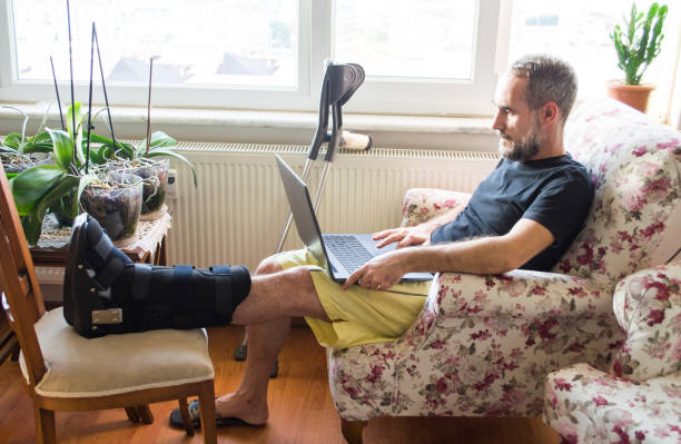Man wearing walking boot for achilles tendon treatment is using laptop at home. Injured man with crutches. Man wearing walking boot for achilles tendon treatment is using laptop at home orthopedic cast stock pictures, royalty-free photos & images
