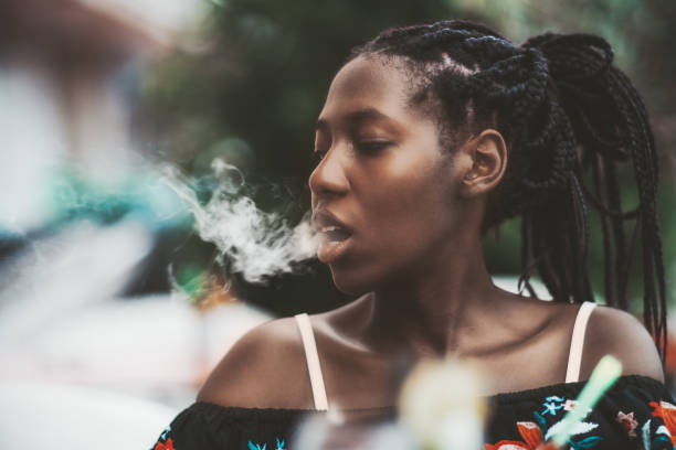Black girl is exhaling hookah smoke Portrait of a dazzling African girl with braids sitting in a street cafe and exhaling smoke from the hookah; young black female outdoors in a bar plays with the vapor from an electronic cigarette weaving photos stock pictures, royalty-free photos & images