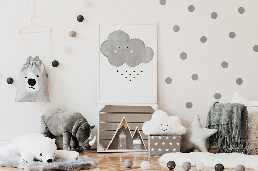 Scandinavian child room with mock up poster frame on the pattern wall, boxes, teddy bear and toys. Cute modern interior of playroom with white walls, wooden accessories and colorful toys.
