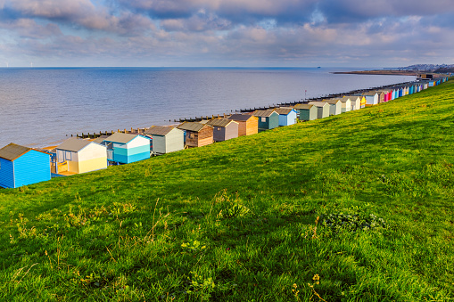 Row of beach huts along the coast in Tankerton, Whitstable, Kent. The green grass slopes are behind the huts and groynes, water breakers can be seen along the beach