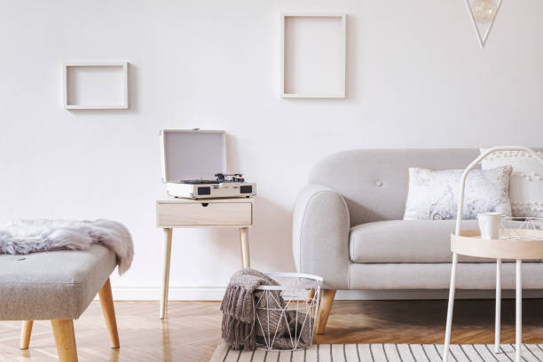 istock Stylish and cozy scandinavian white interior with design sofa, bench, coffee table, pillows, blanket, gramophone and mock up photo frames. White background walls and modern triangle lamp. 1089785278
