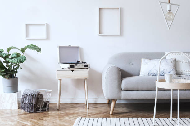 istock Stylish scandinavian white interior with design sofa, tropical plant, pillows, blanket, gramophone and mock up photo frames. White background walls, brown wooden parquet and modern triangle lamp. 1089786676