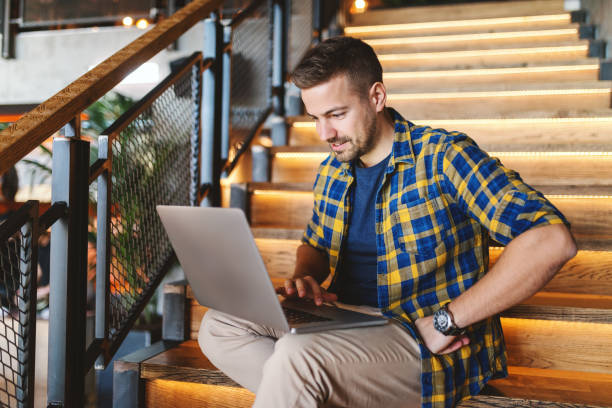istock Smiling bearded freelancer using laptop and sitting on stairs in cafeteria. 1089839964