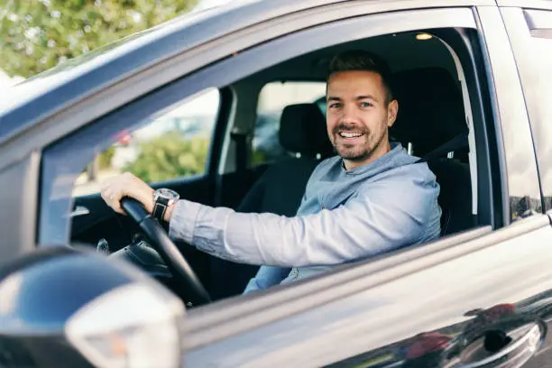 Smiling handsome man looking at camera and driving his car. Hand on steering wheel.
