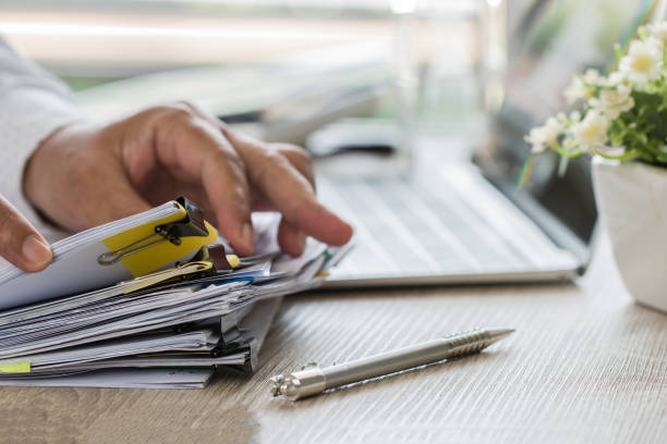 geschäftsmann hände halten stift für die arbeit in stapeln von papierakten suchen informationen geschäftsbericht papiere und haufen von unvollendeten dokumente erreicht auf laptop-computer-schreibtisch im modernen büro - book law legal system lawyer stock-fotos und bilder