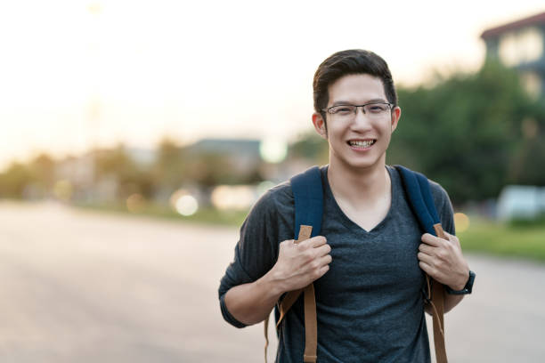 jovem asiática atraente ou universitário segurando a mochila sorrindo para sentimento de câmera animado, boas férias, alegria e positivo com fundo de luz quente do sol em viajar conceito de ásia com espaço de cópia. - tourist backpack smiling cheerful - fotografias e filmes do acervo