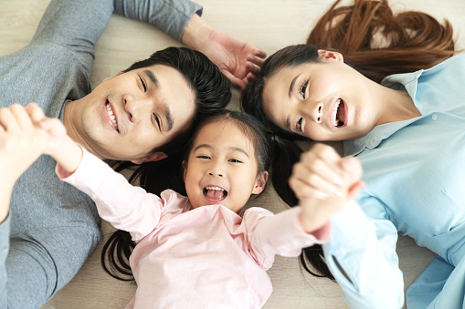 Happy attractive asian family with mom, dad and little cute young daughter or elementary kids holding hands together lying on floor looking at camera on top view feeling enjoy happy time and positive.