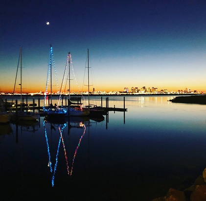 Christmas lights adorn two sailboats in Boston Harbor as the sun sets leaving a beautiful reflection