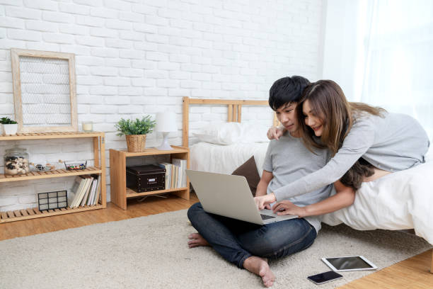 young asian couple looking at laptop computer to search for travel plan, book hotel room, buy ticket, purchase travel insurance or shopping online together at bedroom in e-commerce technology concept. - computer two people asian ethnicity women imagens e fotografias de stock